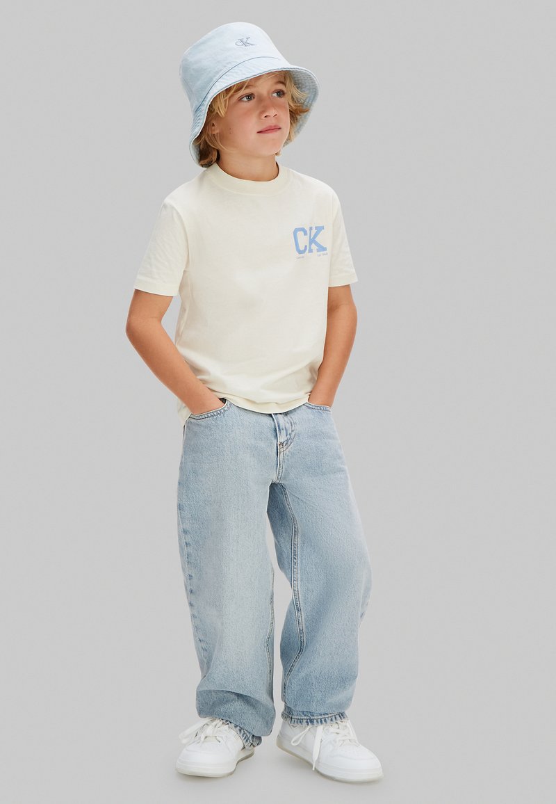 Garçon portant un chapeau bob bleu clair, un t-shirt blanc cassé avec le logo "CK", un jean bleu clair et des baskets blanches, debout les mains dans les poches.