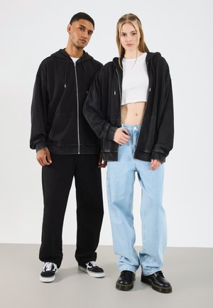 Black zip-up hoodies with kangaroo pockets, over-sized fit. Model 1 wears black pants and black sneakers; Model 2 has light blue wide-leg jeans.