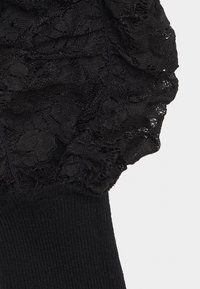 Musta pitsihöyhenhihansuun yksityiskohta teksturoidulla kankaalla, jossa on ribbineulottu musta hihansuu; pitsissä on läpikuultava kukkakuvio, sileä siirtymä kiinteään kangasmateriaaliin.