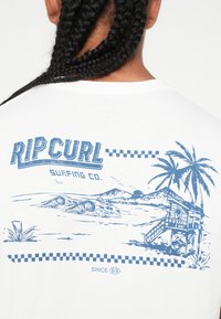 Biała koszulka z niebieskim nadrukiem graficznym przedstawiającym plażę z falami, palmami i wieżą ratunkową, oznakowana napisem "Rip Curl Surfing Co."