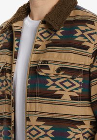 Tan patterned jacket with a brown sherpa collar, featuring geometric designs in brown, green, and purple. Includes front pockets and zipper.