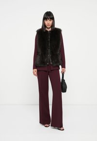 Gilet en fausse fourrure noire, avec des finitions en cuir noir, associé à un blazer ajusté bordeaux et un pantalon large. Sac à main noir, sandales à talons.