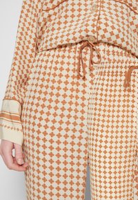 Pantalon beige et rouille à motifs avec une ceinture élastique et un cordon, associé à une chemise boutonnée assortie avec des détails aux poignets.