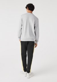 Homme debout, dos à la caméra, portant un sweat-shirt gris clair, un pantalon noir avec des bandes jaunes sur les côtés, et des baskets blanches sur fond blanc.