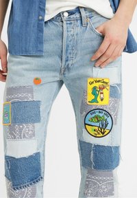 Jeans en un azul claro desgastado que presentan secciones de patchwork, parches bordados y una textura desvaída con múltiples tonos de mezclilla.