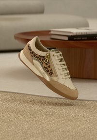 Sneaker in cream leather with leopard print accents and gold details. Features a side zipper, beige suede toe, and a rubber sole.