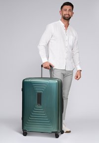 Groene harde koffer met een getextureerd ontwerp met ribbels. Voorzien van een zwart handvat en wielen voor eenvoudige mobiliteit. Merkenlogo zichtbaar.