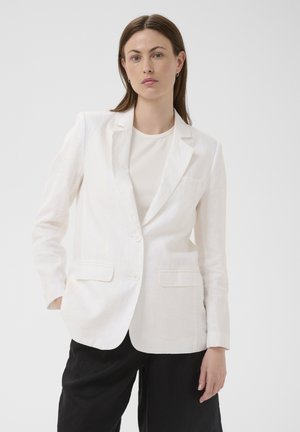 Femme aux cheveux bruns raides portant un blazer blanc et un t-shirt avec un pantalon noir, debout devant un fond clair uni.