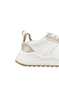 Sneaker bianco con accenti metallici dorati sul tallone, pannello laterale testurizzato e suola spessa. Presenta una linguetta sul retro.