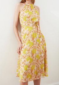 Robe halter florale en pêche clair avec des fleurs jaunes et orange. Elle présente un corsage ajusté, une taille à nouer et une jupe fluide. Texture de tissu lisse.