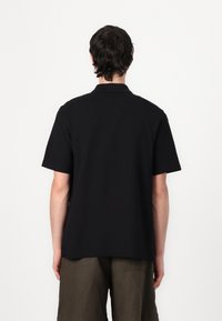 Cămașă polo neagră cu mâneci scurte, guler clasic și tiv drept. Materialul pare neted. Se potrivește cu pantaloni scurți cremoși.