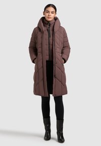 Abrigo largo acolchado marrón con collar alto, superpuesto a un abrigo acolchado más corto, con cremallera frontal y bolsillos laterales. Leggings negros y botas.