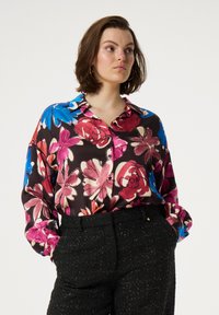Bloemige zijden blouse met een donkere achtergrond, versierd met grote roze, rode en blauwe bloemen. Zachte textuur en een knoopsluiting aan de voorkant.