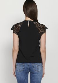 Zwart top met kant cap sleeves en een keyhole detail op de rug, met een gladde lijfje en intrigerende kant textuur op de schouders. Gecombineerd met blauwe jeans.