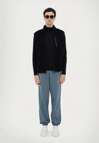 Melns vilnas veste ar rāvējslēdzēju pār melnu augstu apkakli, kopā ar gaiši zilām svārku biksēm ar nelielu daudzkrāsainu logotipu un baltiem sporta apaviem.