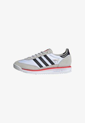 Hvit sneaker med grå semsket skinn detaljer, svarte striper, røde detaljer og en teksturert gummisåle. Har polstret krage og lisser.