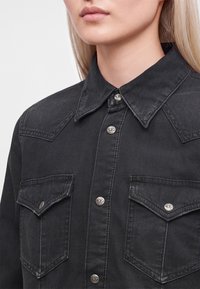 Gros plan d'une personne portant une chemise en denim noire avec des boutons-pression métalliques et des poches à rabat sur la poitrine.