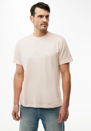 Hombre con cabello corto y oscuro, vistiendo una camiseta de manga corta rosa claro y vaqueros azules, mirando hacia la derecha sobre un fondo blanco.