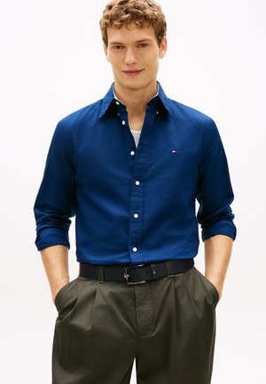 Mladić s kovrčavom kosom koji nosi plavu košulju na kopčanje, tamno zelene hlače i crni remen, stoji s rukama u džepovima.