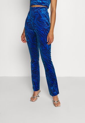 Vrouw draagt blauwe broek met hoge taille en donkere, draaiende patronen, gecombineerd met zilveren, openhiel sandaal met hak.