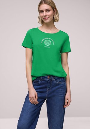 Femme portant un T-shirt vert avec l'inscription « Summer is my season Côte d'Azur » et un motif coquillage, associée à un jean bleu, debout devant un fond uni.