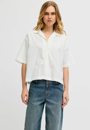 Mujer con cabello rubio hasta los hombros que lleva una camisa blanca de manga corta y botones suelta y vaqueros azules de denim sobre un fondo liso.