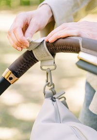 Witte tas bevestigd aan een zwarte handgreep met een gouden accent. Een grijze riem bevestigt een gedeelte van de bruine geverniste grip, met zichtbare stiksels.