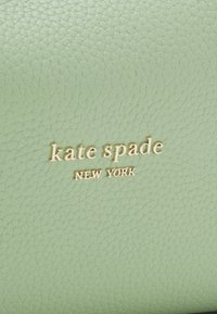 Zielona teksturowana skóra z wytłoczonym złotym logo "kate spade NEW YORK". Powierzchnia wydaje się gładka z lekko błyszczącym wykończeniem.