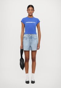 T-shirt in cotone blu con la scritta "PRIORITY" in bianco, abbinata a pantaloni corti di denim chiaro. La modella tiene una borsa nera, indossa calze bianche e scarpe nere.