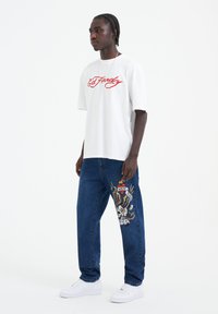 White cotton t-shirt with red graphic text, paired with blue denim jeans featuring a colorful embroidered design on the left leg. White sneakers.