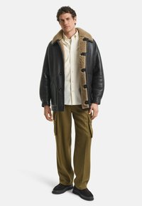 Schwarzer Ledercoat mit einem beigen, pelzigen Kragen, Knopfverschlüssen, kombiniert mit olivgrünen Cargo-Hosen und schwarzen Schuhen. Das Model steht vor einem weißen Hintergrund.