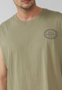 T-shirt sans manches vert olive en coton doux, avec un col rond et un logo imprimé circulaire sur la zone gauche de la poitrine.