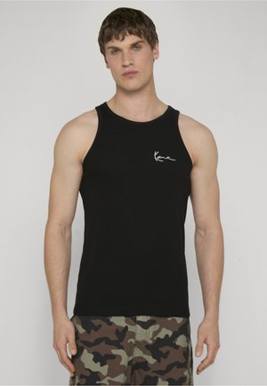 Hombre con camiseta negra sin mangas con logo blanco en el pecho y pantalones cortos de camuflaje verde y marrón, de pie frente a un fondo liso.