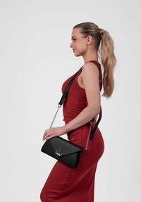Černá kožená křížová taška s trojúhelníkovým klopou, stříbrným logem, s řetízkovým popruhem. Modelka má na sobě červené upnuté šaty.