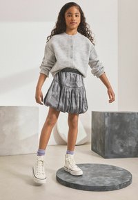 Grauer gestrickter Pullover mit drei Knöpfen, kombiniert mit einem glänzenden grauen Plisseerock. Weiße High-Top-Sneaker und lilafarbene Socken runden das Outfit ab.