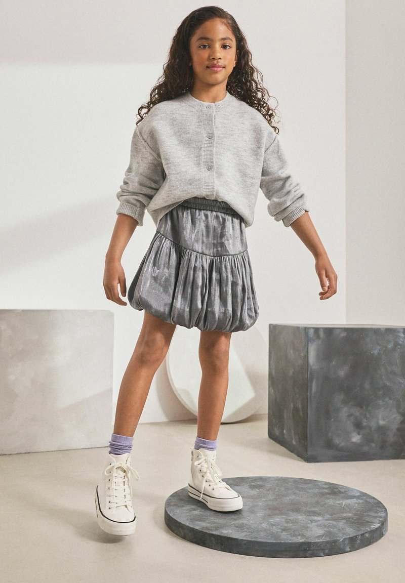 Grauer gestrickter Pullover mit drei Knöpfen, kombiniert mit einem glänzenden grauen Plisseerock. Weiße High-Top-Sneaker und lilafarbene Socken runden das Outfit ab.