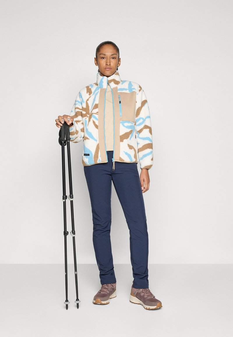 Kobieta w wzorzystej polarowej kurtce i ciemnych spodniach stoi z dwoma czarnymi kijami trekkingowymi na jednolitym tle.