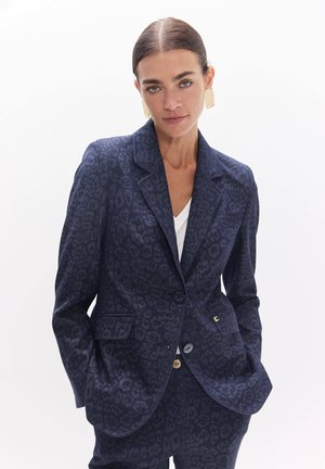 Mujer con el cabello recogido hacia atrás que lleva pendientes rectangulares dorados y una americana azul oscuro con estampado de leopardo, pantalones a juego y top blanco.