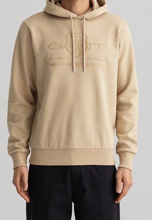 Mand iført beige hættetrøje med GANT 1949 American Sportswear broderet logo, kombineret med sorte bukser, mod ensfarvet baggrund.