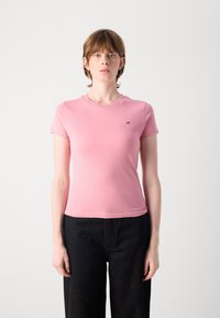 T-shirt en coton rose avec un col rond et des manches courtes, arborant un petit logo sur la poitrine, associé à un pantalon noir taille haute.