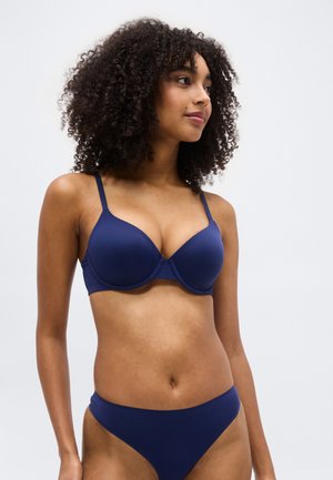 Žena s kučeravými vlasmi, oblečená v zodpovedajúcom tmavomodrom podprsenkovom a spodnom prádle, mierne otočená doprava na jednoduchom bielom pozadí.