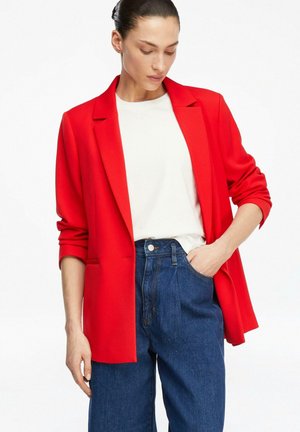 Kobieta w jasnoczerwonej marynarce na białej koszuli i niebieskich dżinsach z wysokim stanem, z jedną ręką w kieszeni, patrząca w dół.