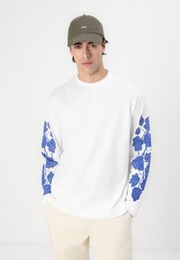 Witte langwoning overhemd met blauwe bloemenprint op de mouwen, gecombineerd met een khaki baseballpet met geborduurd logo en lichtgekleurde broek.