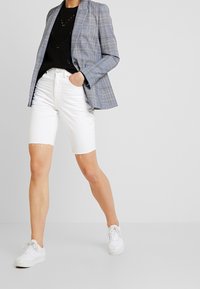 Eine Person steht selbstbewusst in einem karierten Blazer, einem schwarzen Stricktop und weißen Jeansshorts, kombiniert mit weißen Sneakers, vor einem schlichten Hintergrund.