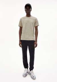 Khaki T-shirt met korte mouwen in combinatie met zwarte joggers; het model draagt grijze sneakers en een zwarte pet met een wit embleem. Eenvoudig, casual ontwerp.