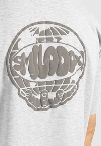 Camiseta gris de manga corta con un gran gráfico en relieve de manos sosteniendo un globo, acompañado de un texto estilizado "SMILODX". Textura de tela suave y casual.
