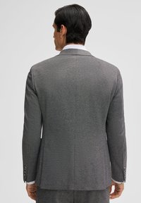 Giacca da suit grigia con tessuto testurizzato, spacco singolo, revers ad angolo, e polsini con tre bottoni, cuciture visibili lungo le cuciture.