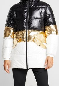 Pohodlná bunda v černé, bílé a zlaté barvě. Má lesklou tkaninu, kapuci, zipový uzávěr a elastické manžety. Obsahuje boční kapsy a design s barevnými bloky.