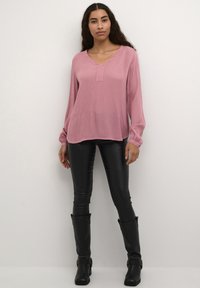 Helles rosa langärmliges Bluse mit V-Ausschnitt und mittigem Knopfverschluss, kombiniert mit engen schwarzen Lederhosen und kniehohen schwarzen Stiefeln.