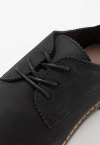 Chaussure en cuir noir avec des bords festonnés, ornée de lacets noirs et d'une semelle cousue. Texture lisse, design minimaliste.